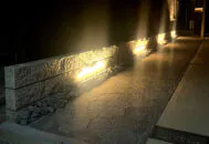 照明で照らされた夜のアプローチ。ダークカラーのインターロッキングブロック、フローで舗装。塀には白い割肌と黒の種石が特徴のダルメを使用。塀の足元を天然石のアウトドアロックが彩る。