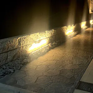 照明で照らされた夜のアプローチ。ダークカラーのインターロッキングブロック、フローで舗装。塀には白い割肌と黒の種石が特徴のダルメを使用。塀の足元を天然石のアウトドアロックが彩る。